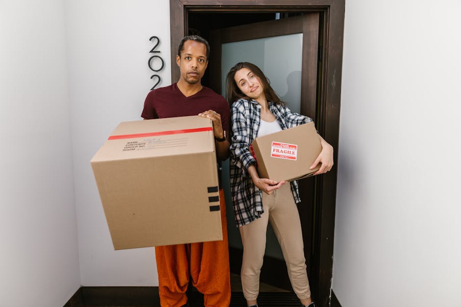 A man and a woman standing at a doorway inside a property during a home relocation, both holding cardboard boxes with packing labels; the man, wearing a maroon t-shirt and orange pants, holds a larger