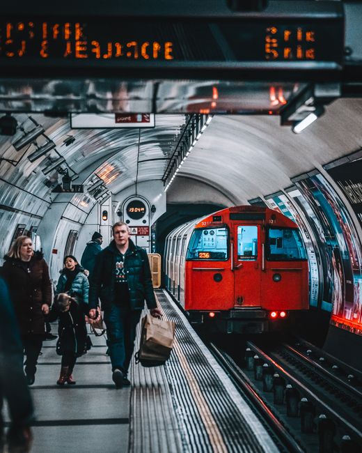 Inside a London underground station with a curved, tiled ceiling and illuminated signage overhead displaying station information. Several passengers are on the platform, including a man carrying a large paper shopping bag, a woman and a young girl walking together, all dressed in casual clothing suitable for urban travel. In the background, a red London Underground train is either arriving or departing, with its doors closed and a digital display showing the train’s destination. The platform surface has tactile paving along the edge and is equipped with safety features such as handrails and lighting. The station environment reflects a typical urban setting for home relocation or furniture transport, with an emphasis on passenger movement and transit logistics, aligned with moving services offered by Balham Man and Van.