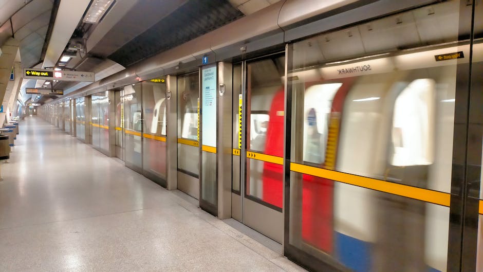 The image depicts an empty platform at Balham station with a row of closed glass doors and sliding doors separating the platform from the train tracks. The platform surface is smooth and clean, made of light-colored material, with minimal clutter. Overhead signage indicates directions such as 'Way out' and provides information relevant to visitors. In the background, a train with a distinctive red and white exterior is visible through the glass doors, which are slightly reflective and show a blurred view of the train interior and station environment. The area is illuminated with ceiling-mounted lighting, creating a bright, neutral setting suitable for passengers heading to or arriving from train travel. This setting exemplifies a typical station environment, associated with transportation and logistics, supporting activities like home relocation or furniture transport when planning removals with local services such as Balham Man and Van.