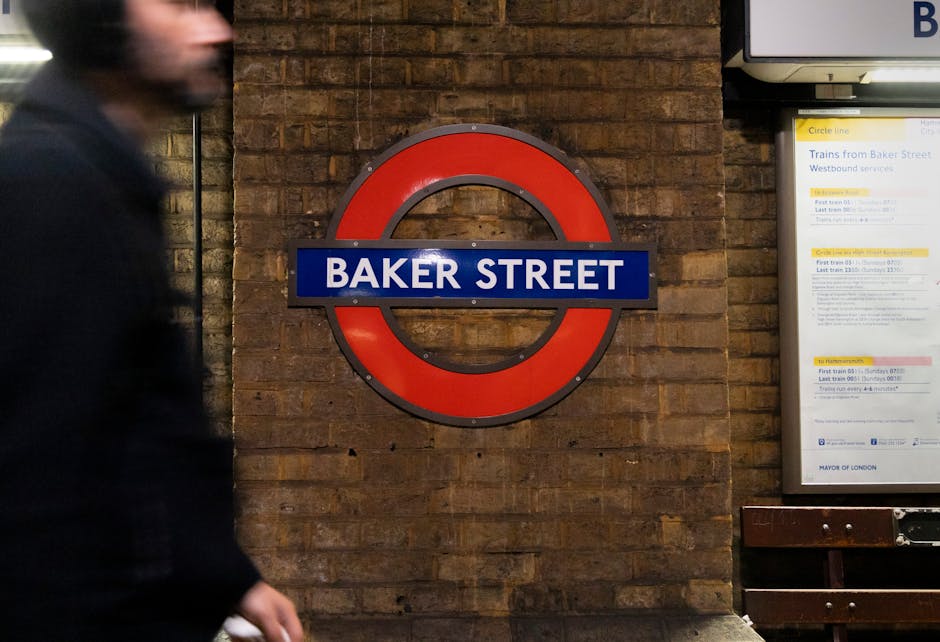 Inside a London underground station with a curved, tiled ceiling and illuminated signage overhead displaying station information. Several passengers are on the platform, including a man carrying a large paper shopping bag, a woman and a young girl walking together, all dressed in casual clothing suitable for urban travel. In the background, a red London Underground train is either arriving or departing, with its doors closed and a digital display showing the train’s destination. The platform surface has tactile paving along the edge and is equipped with safety features such as handrails and lighting. The station environment reflects a typical urban setting for home relocation or furniture transport, with an emphasis on passenger movement and transit logistics, aligned with moving services offered by Balham Man and Van.