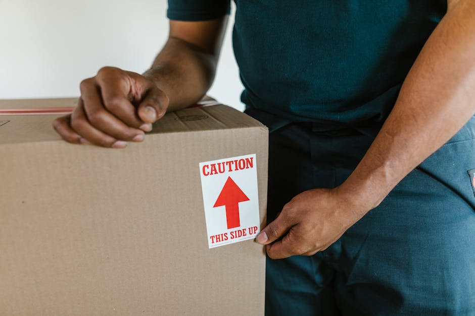 A person wearing a dark green shirt and dark trousers is sealing a large cardboard box with a red and white caution label that reads 'Caution: This Side Up,' featuring an upward-pointing arrow. The individual is indoors, standing next to a packed moving van or loading area, where other boxes, wrapped furniture, and packing materials such as bubble wrap and plastic wrapping are visible. The scene captures the process of packing and preparing household items for home relocation, with the focus on proper handling and safety measures during furniture transport and loading. The background shows a plain, light-colored wall, and the lighting is natural, highlighting the careful sealing of the box to prevent damage during transit, illustrating typical steps involved in professional removals carried out by Balham Man and Van.